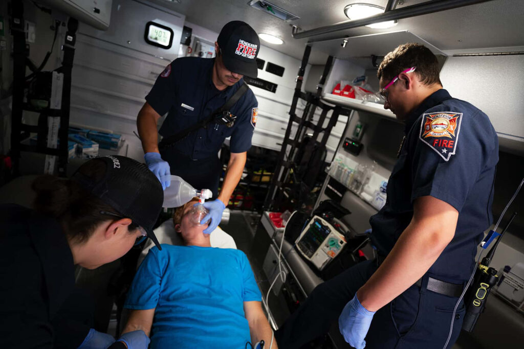 Three emergency responders in uniform treat a person lying on a stretcher inside an ambulance, using medical equipment and wearing gloves.