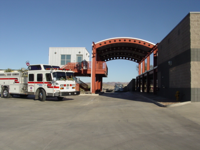 A fire truck is parked outside a modern fire station with a large arched red metal canopy and attached gray building on a sunny day. Mountains are visible in the background.